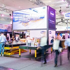 The DEMMELER stand at EuroBLECH 2026 with the LED wall "One line. Any length." and 3D welding tables, visitors blurred in motion in front of it